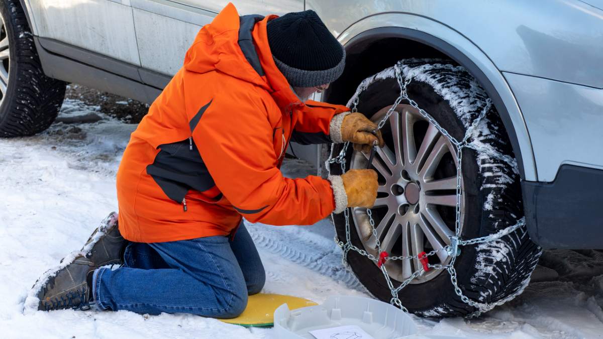 A man in a n orange snow jacket placing snow chains on a car wheel 