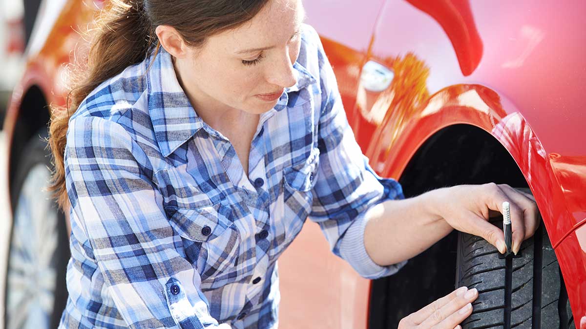 Woman in blue checked shirt checking tyre tread depth on red car using gauge