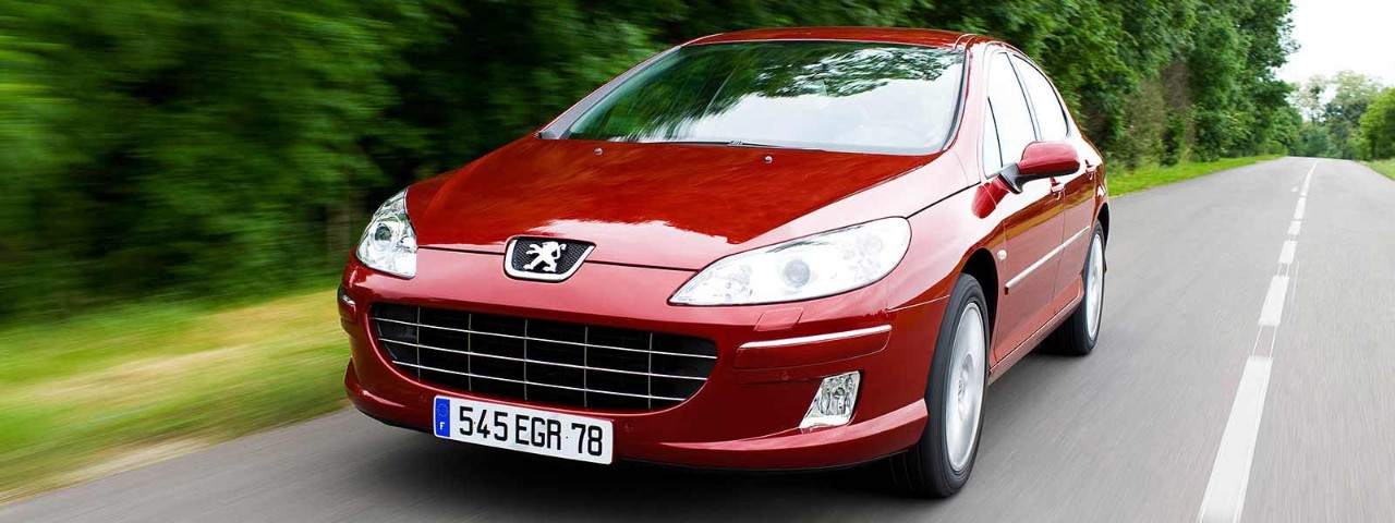 Red car driving along country tree-lined road.