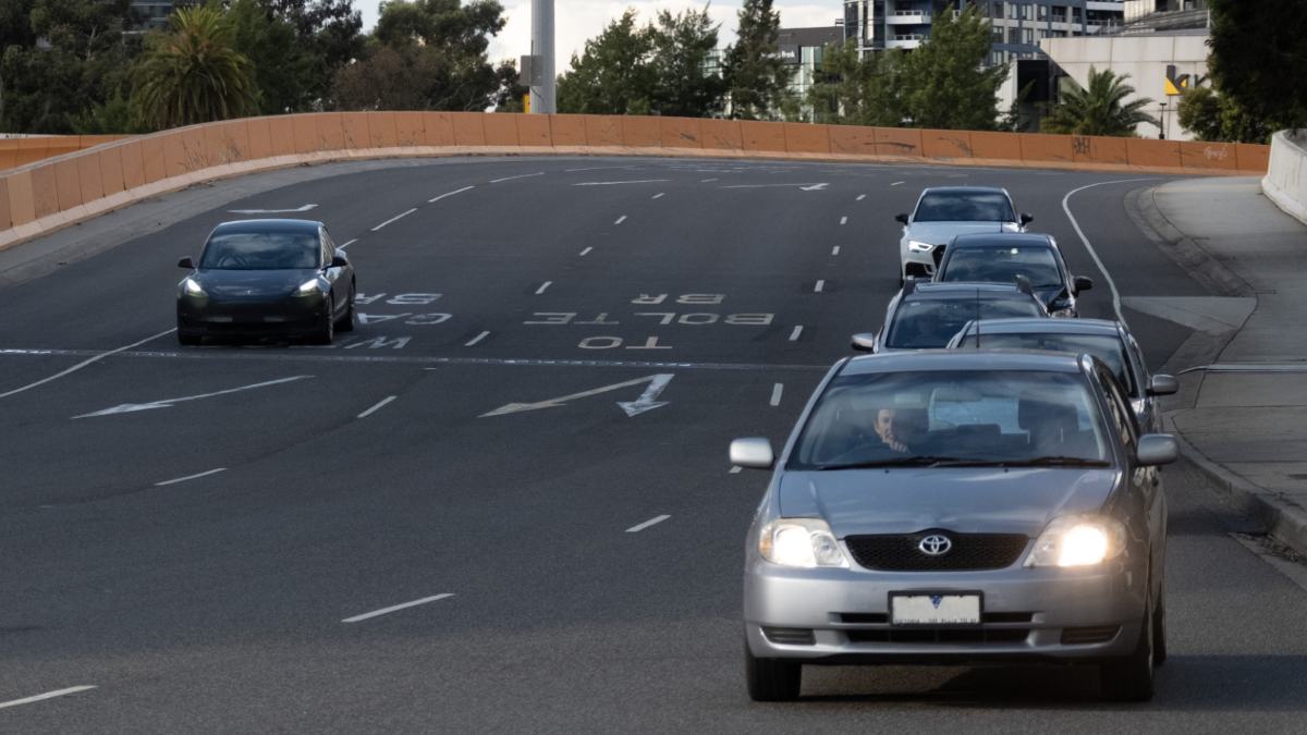 Several cars driving on a four-lane urban road in Melbourne during the day. Several of the cars have daytime running lights (DLRs) or their low beam headlights on.