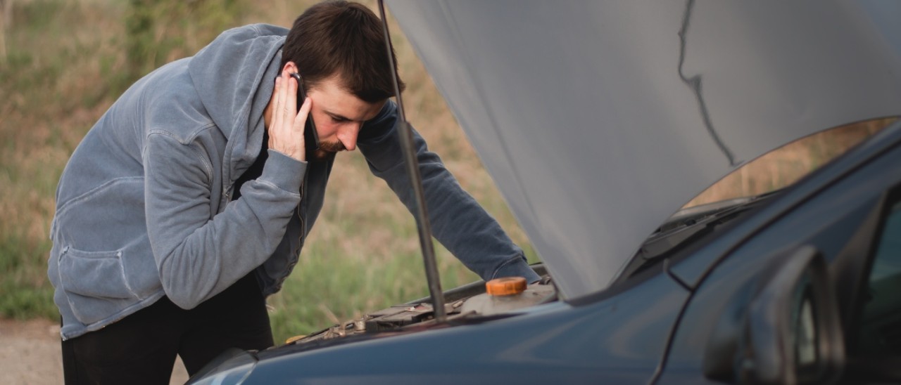 A man holding a phone up to his ear while he looks under the bonnet of his car, which is parked on the side of the road, near a field