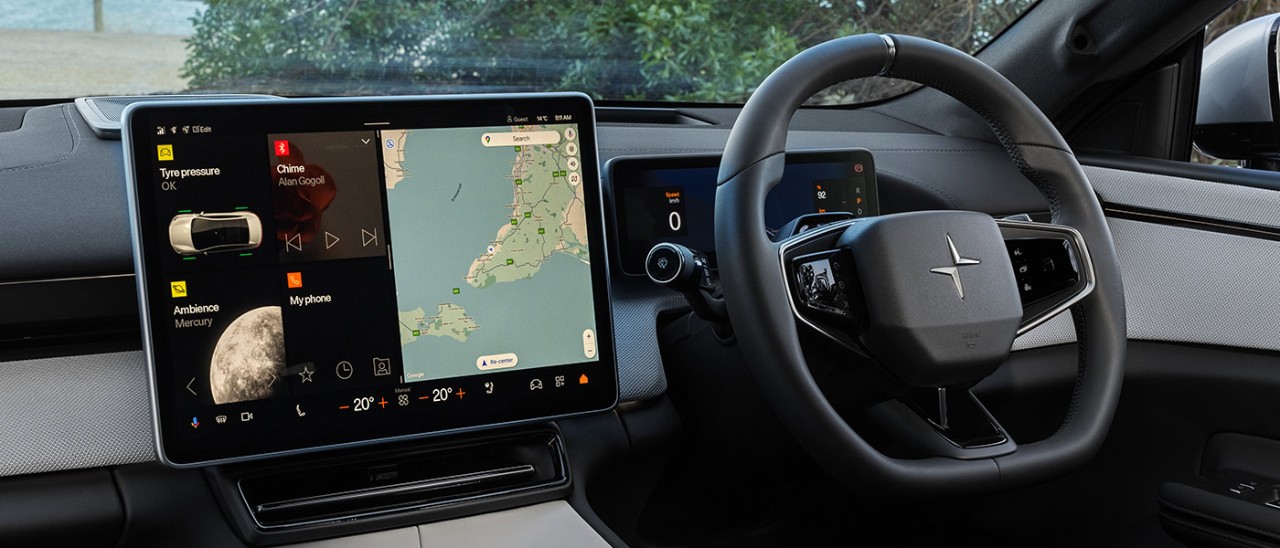 2025-Polestar-4-SUV-dash-banner-1400x600 Interior of Polestar 4 prestige SUV at beachside showing large central touchscreen and steering wheel.