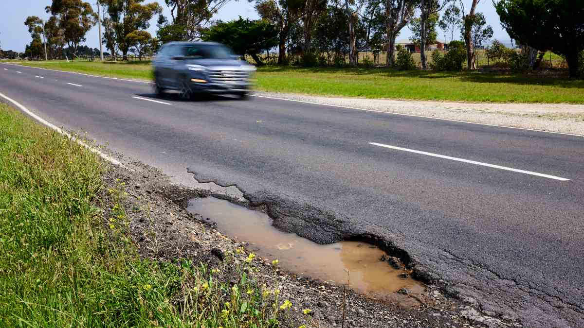 car driving on far side of road; on near side of road, a deep wet pothole