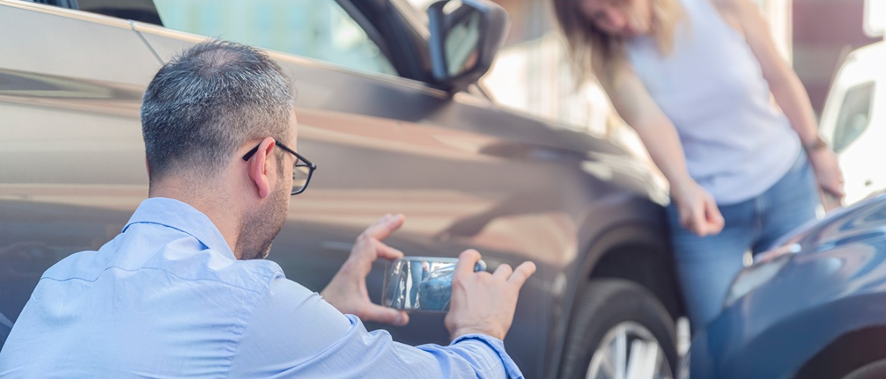 Man bends down to photograph woman pointing to damage to two cars involved in minor crash.