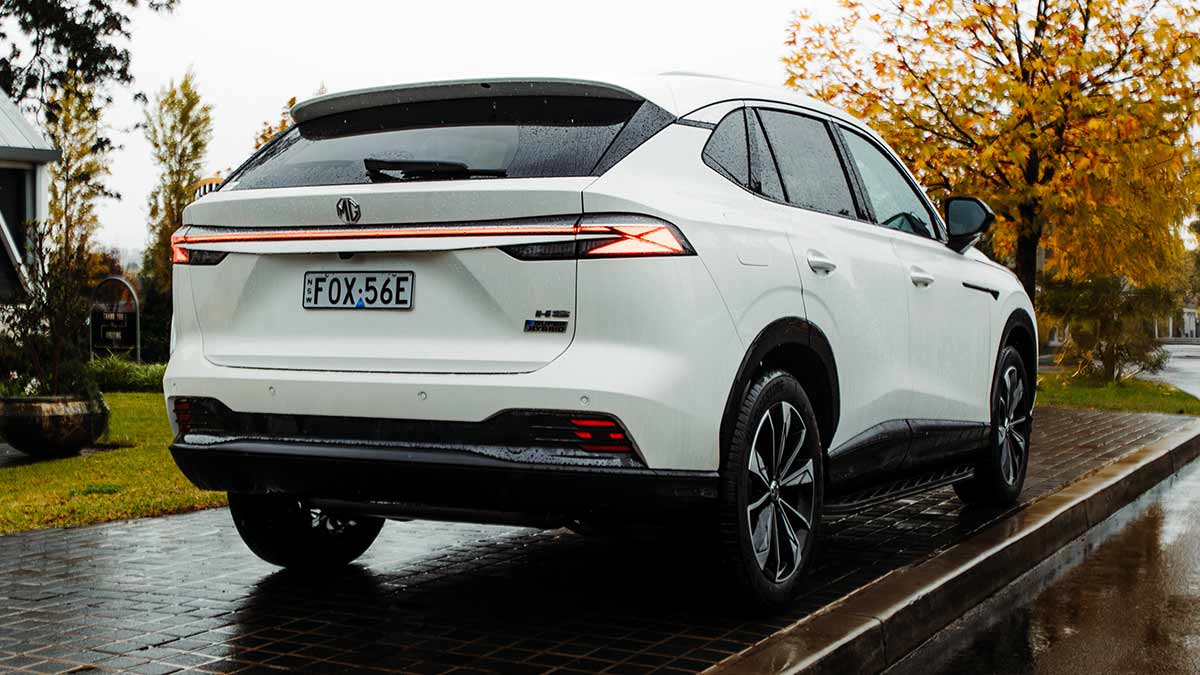 Rear view of white MG HS medium-size SUV on wet pavement in front of tree with yellow autumn leaves