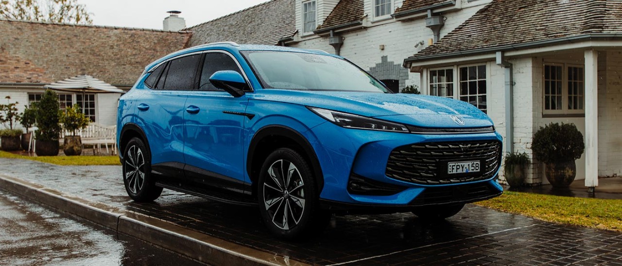 2025-MG-HS-Super-Hybrid-Excite-banner-1400x600 Blue MG HS medium-size hybrid SUV on wet pavement in front of white-brick heritage house.