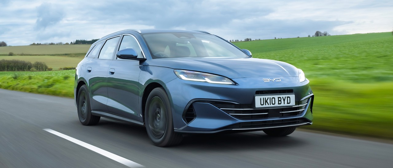 Blue metallic BYD Seal 6 plug-in hybrid station wagon driving through lush green countryside in England.