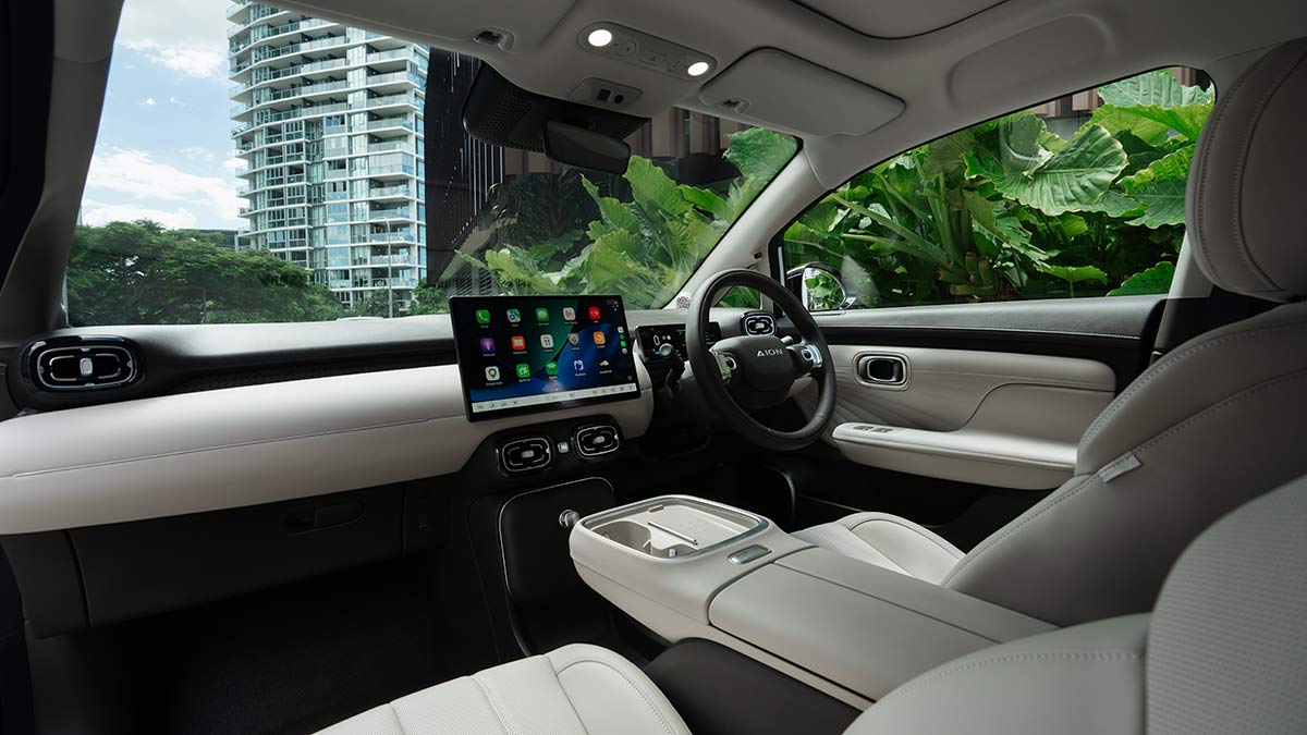 View of GAC Aion UT cockpit from rear left passenger seat, showing long centre console, cream/charcoal trim and large centre touchscreen.