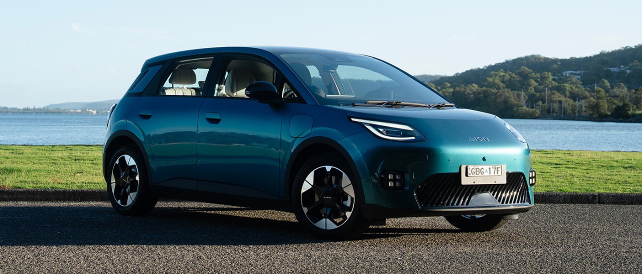 Green GAC Aion UT electric hatch at waterfront carpark with bay and bushland in background.