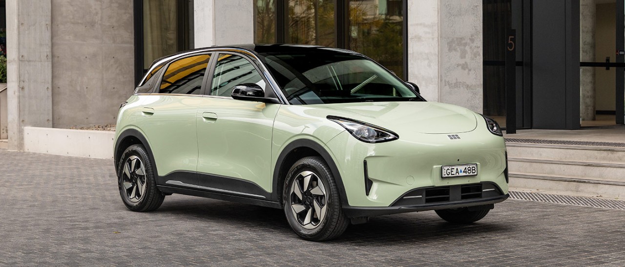 Light green Geely EX2 electric hatch with black roof, parked on grey paved area outside city building with thick concrete pillars.