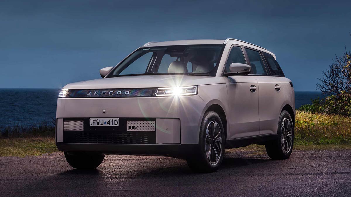 White Jaecoo J5 EV small SUV in carpark fronting coastal shrub and ocean at dusk with slim LED headlights on.