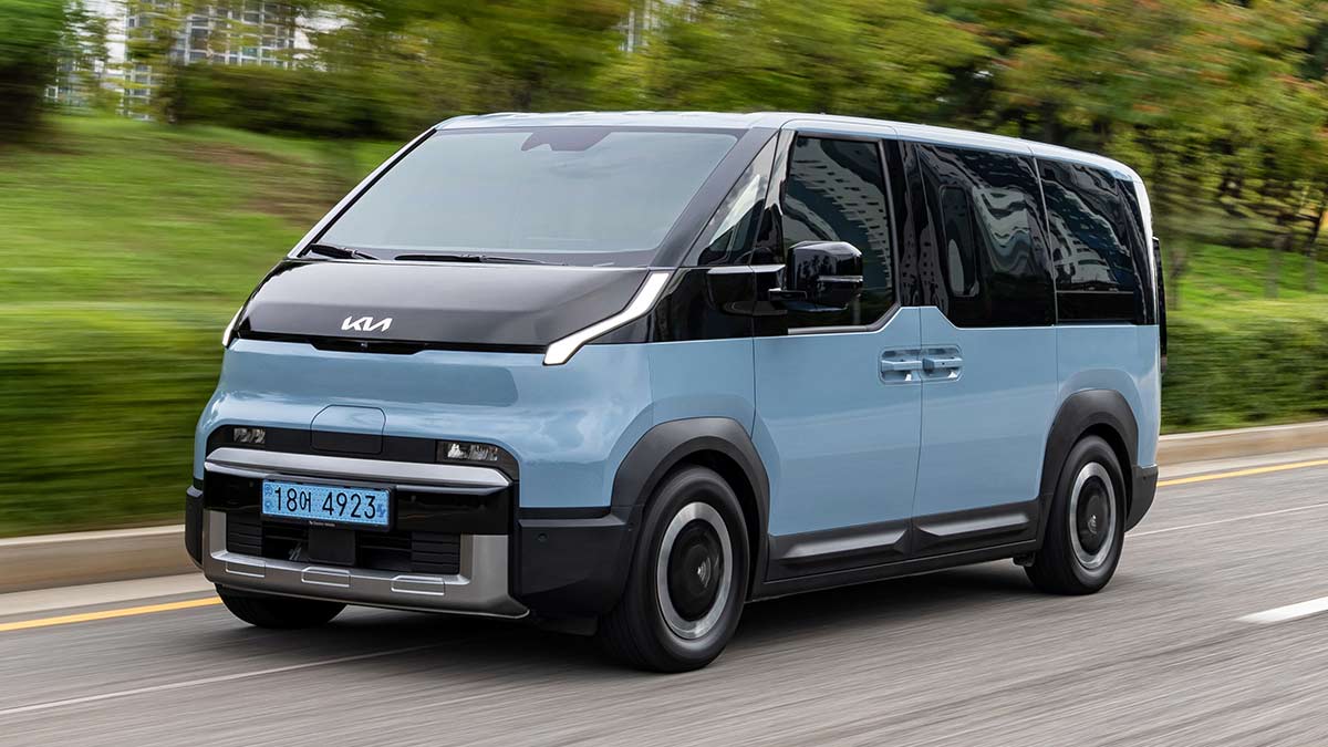 Blue Kia PV5 Passenger electric people-mover on road in South Korea with high green bank and buildings behind.