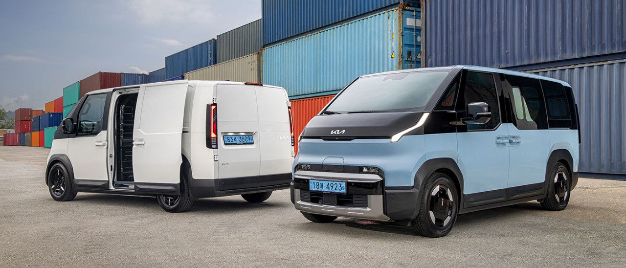 Blue Kia PV5 Passenger EV people-mover and white PV5 Cargo van in front of triple-stacked coloured shipping containers.