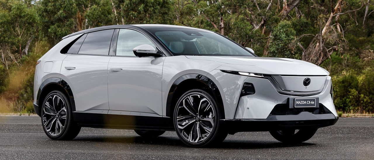 White LHD Euro-spec Mazda CX-6e SUV on forecourt with native bush behind at Lang Lang proving ground in Victoria.