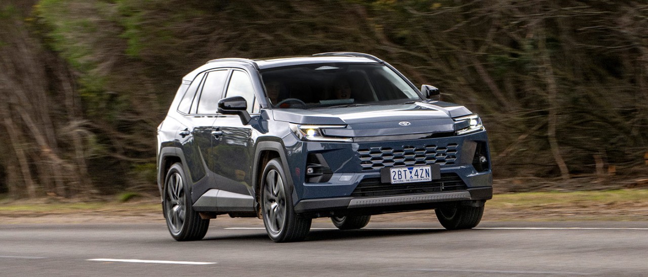 Metallic blue Toyota RAV4 SUV driving on country road with green verge and thick native scrub behind.