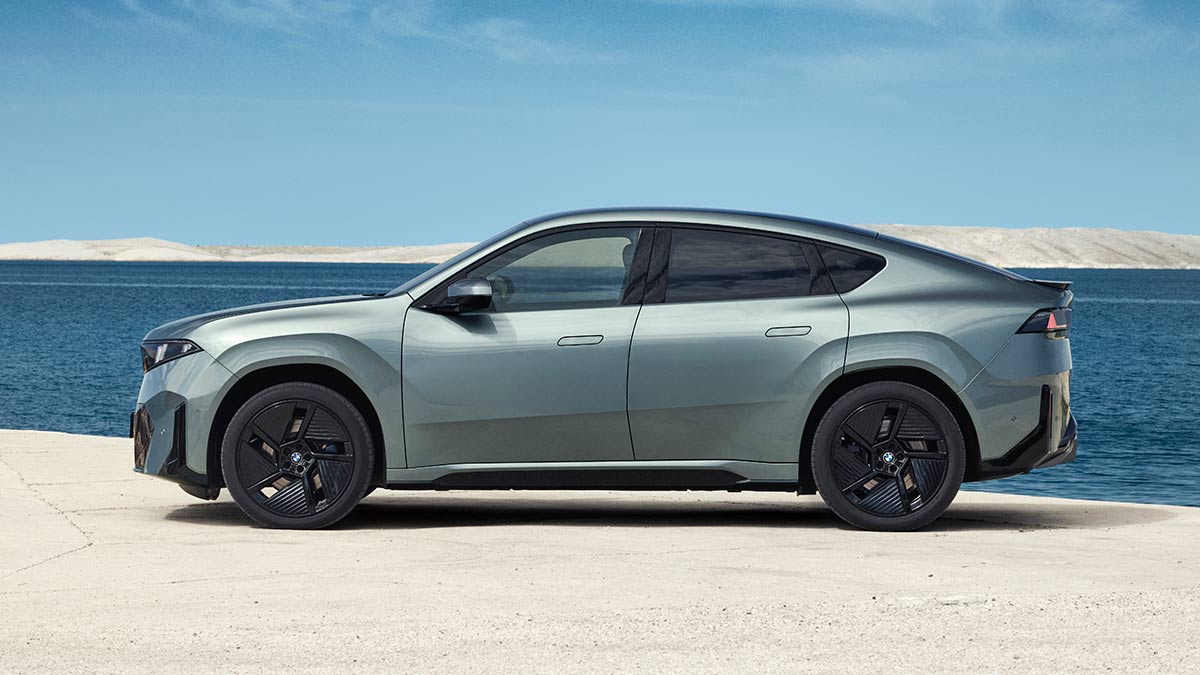 Profile view of metallic sage green BMW iX4 50 xDrive coupe-style SUV in left-hand-drive European-spec on beachfront.