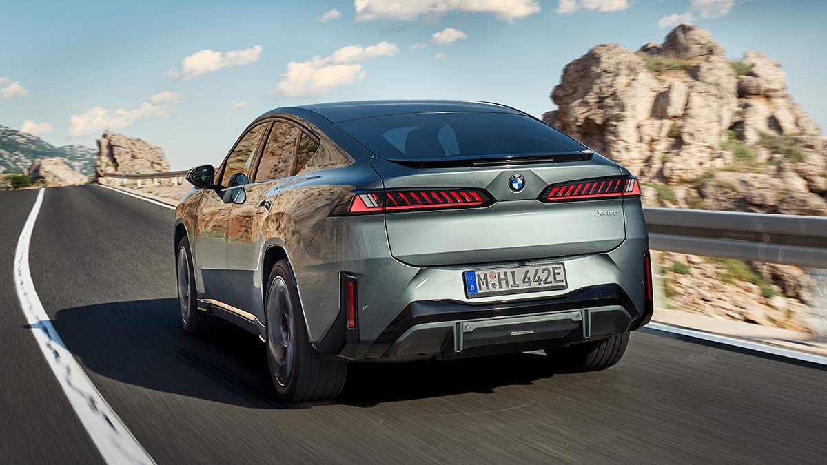 Rear view of metallic sage green BMW iX4 50 xDrive coupe-style SUV in left-hand-drive on coastal road in Europe.