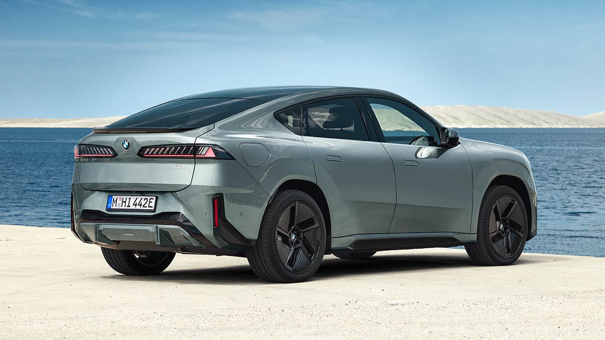 Rear view of metallic sage green BMW iX4 50 xDrive coupe-style SUV in left-hand-drive European-spec on beachfront.