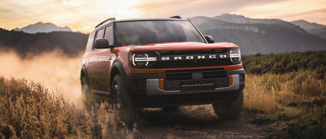 Red Ford Bronco Basecamp plug-in hybrid SUV kicking up dust along narrow grass-lined track in high country.