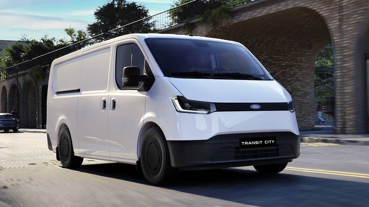 White European-spec Ford Transit City electric van driving on road next to high-walled brick overpass. 