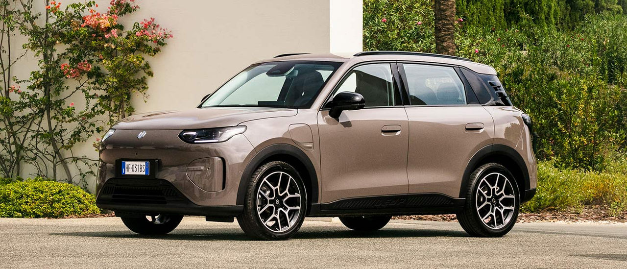 Light brown Leapmotor B03X small electric SUV parked on driveway in outdoor garden setting with thick green shrubbery and white wall with pink rosebush.