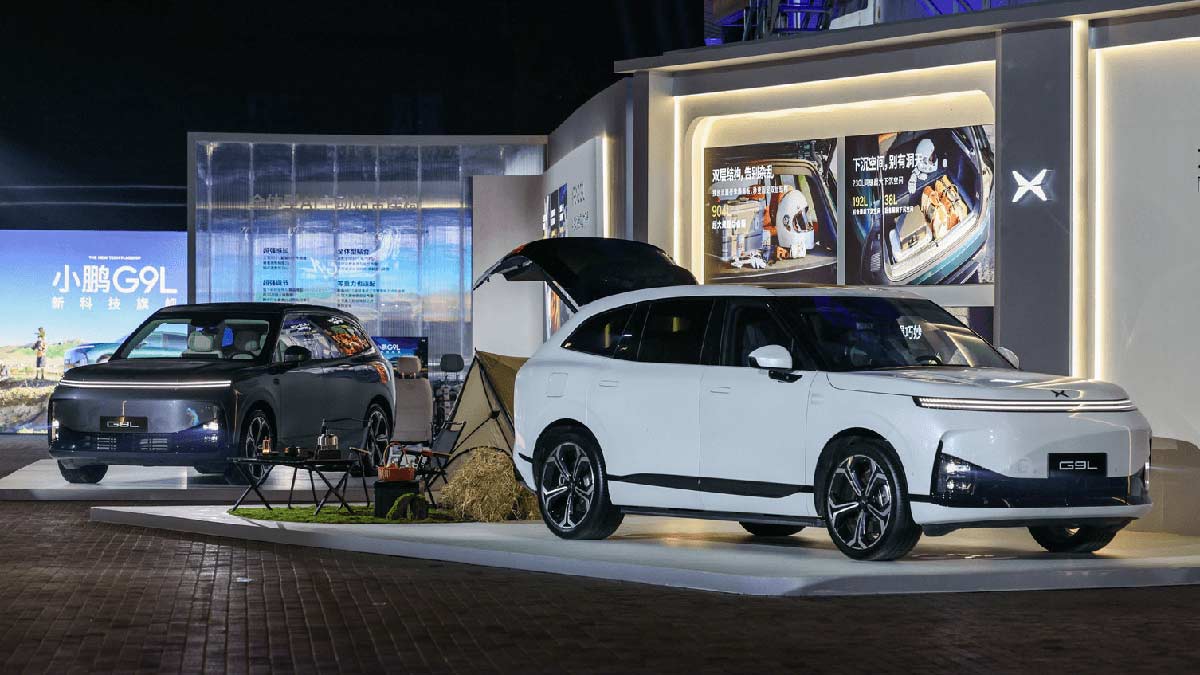 Two XPeng G9L large SUVs on stand at motor show in China, one grey with bright full-width twin LED light bar and the other white with tailgate open.