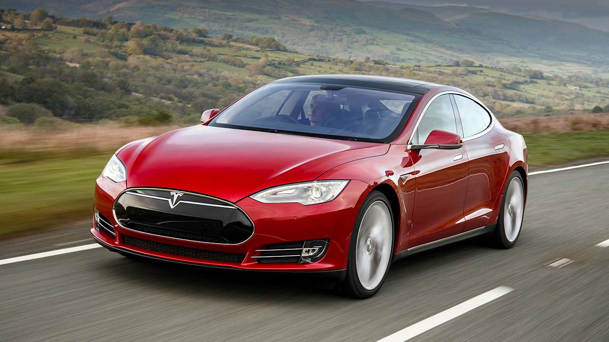 Red 2014 Tesla Model S electric luxury sedan driving on elevated rural bitumen road with mountain ranges in distance.