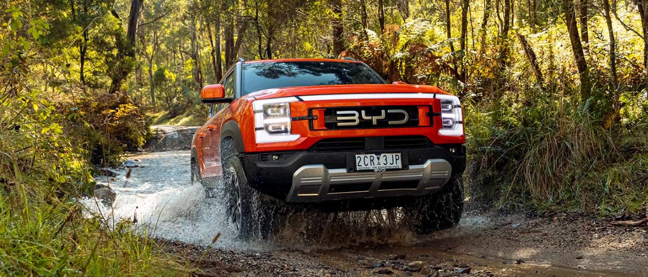 Orange BYD Shark 6 Performance ute driving through water crossing on muddy and rocky 4WD track in sunlit bushland location. 