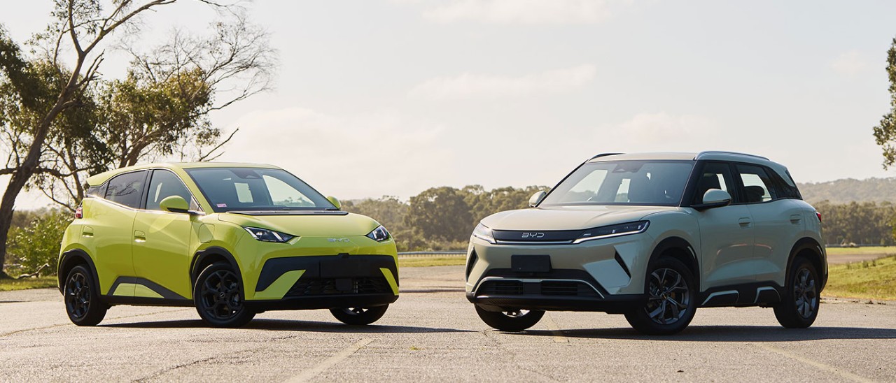 Yellow BYD Atto 1 hatch and cream-coloured BYD Atto 2 SUV parked at Lang Lang proving ground with gum trees behind. 