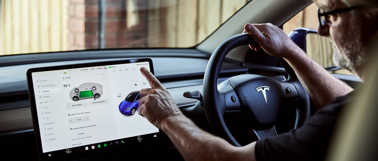 Tesla-Model-Y-EV-home-charge-RACV-7346-banner-1400x600 Bearded man with glasses behind wheel of Tesla Model Y in carport checking EV charge status on touchscreen.