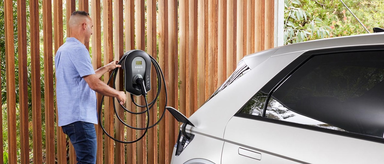 RACV-EV-home-charge-0960-banner-1400x600 Man in blue shirt in carport getting connector from home EV charger on wall for white Hyundai Ioniq 5 EV in foreground