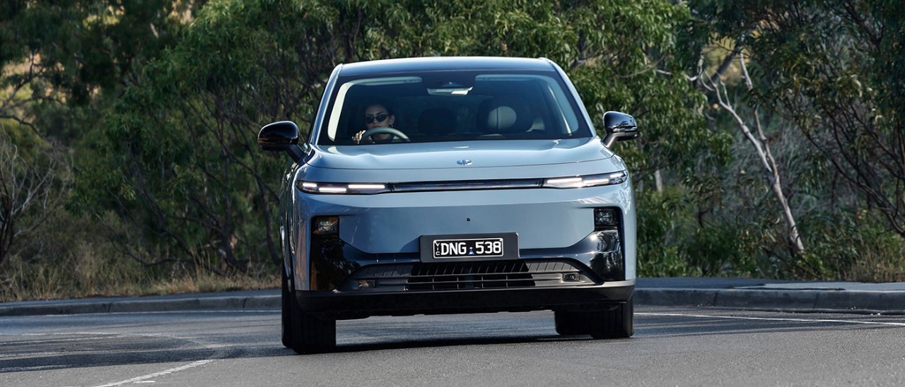 Woman with sunglasses driving blue Leapmotor B10 small electric SUV around corner on tree-lined country road.