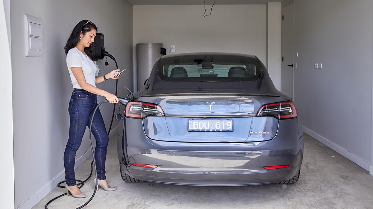 Woman in white shirt and blue jeans charging blue Tesla EV in suburban garage