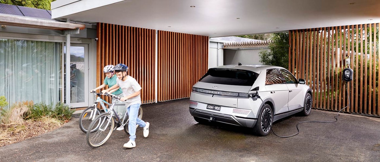 RACV-EV-home-charger-1354-banner-1400x600 White Hyundai Ioniq 5 EV being charged in carport of suburban house next to two children with bicycles