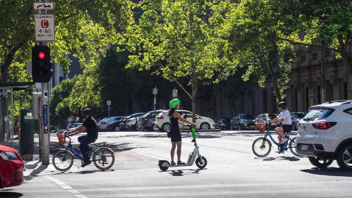 A person riding an e-scooter through a Melbourne CBD intersection. Cars and two cyclists are also passing through the intersection.