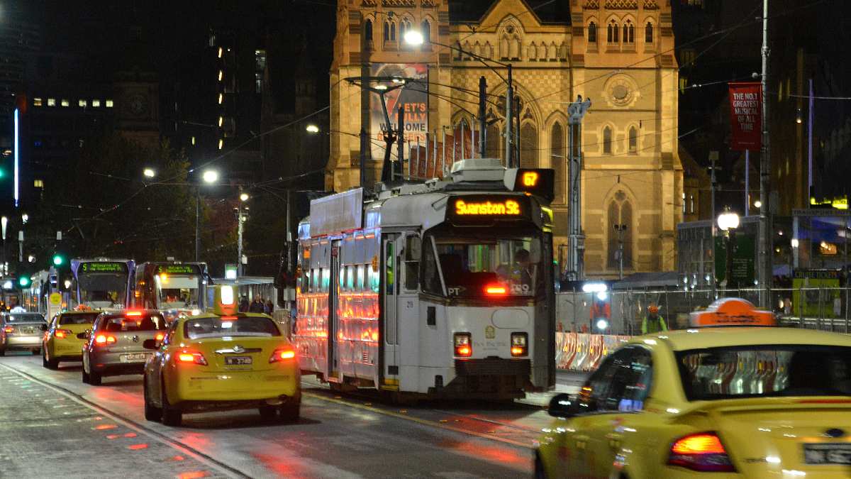 Taxis and cars on a road in inner city Melbourne next to a tram at night time