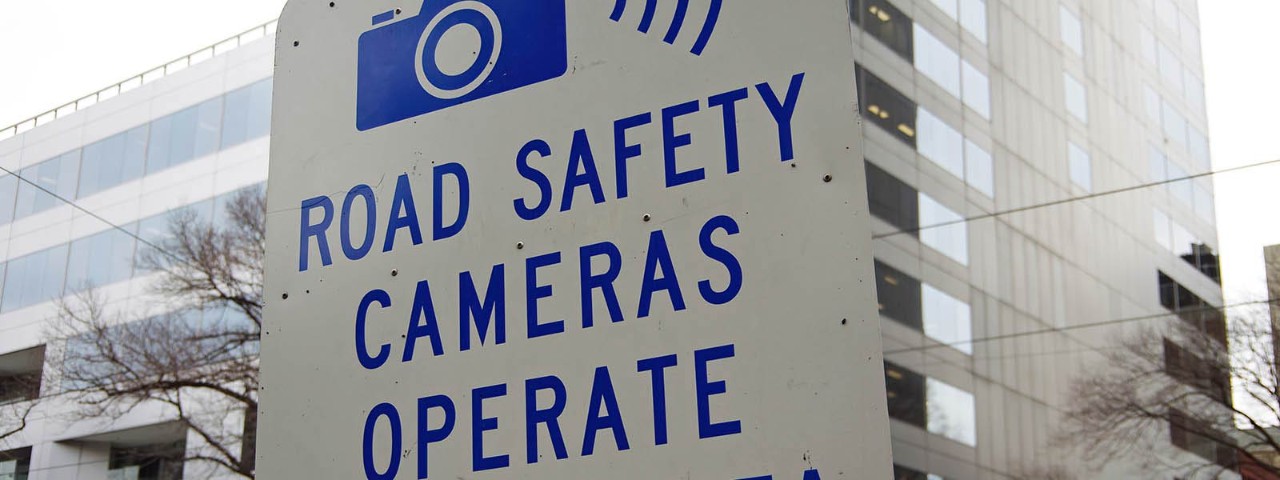 Road safety camera sign with building in the background.