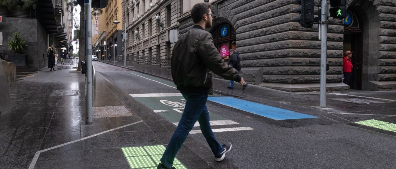 A man walking across a pedestrian crossing at the intersection of Swanston and Little Collins streets in Melbourne. It has been raining and the crossing features green light up tactile markers 