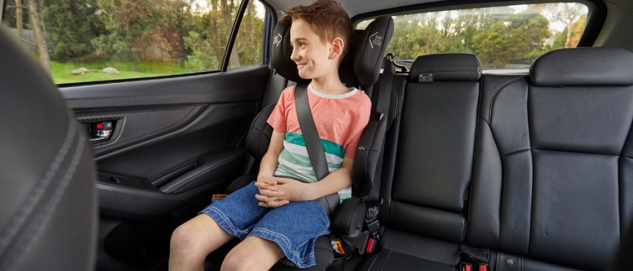 A young school-aged boy sits smiling in a booster seat secured in a car's back seats