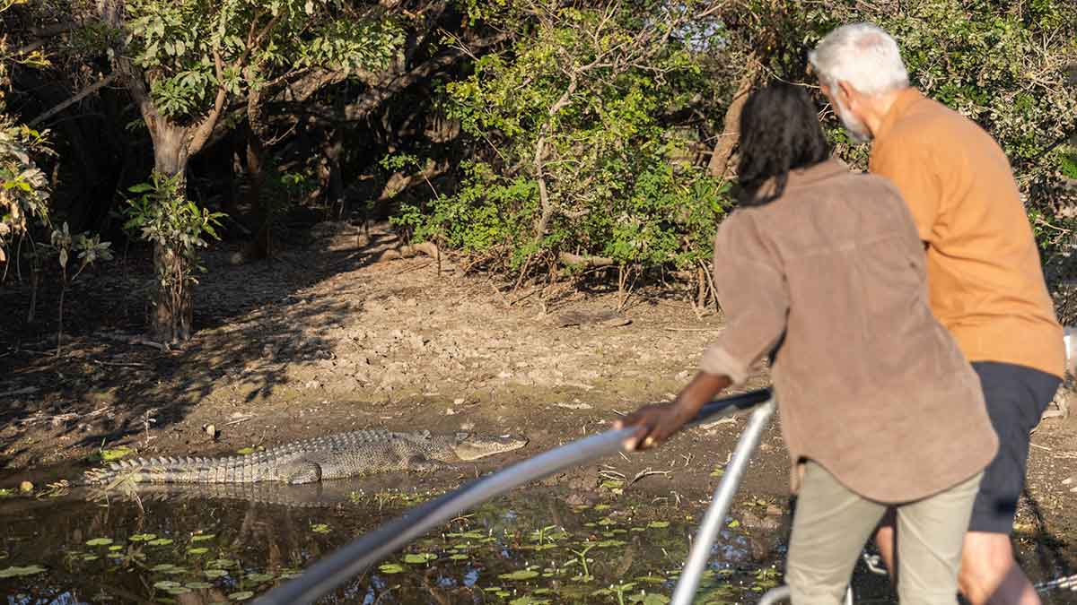 Two people holding on to boat rail looking at crocodile. on river bank.