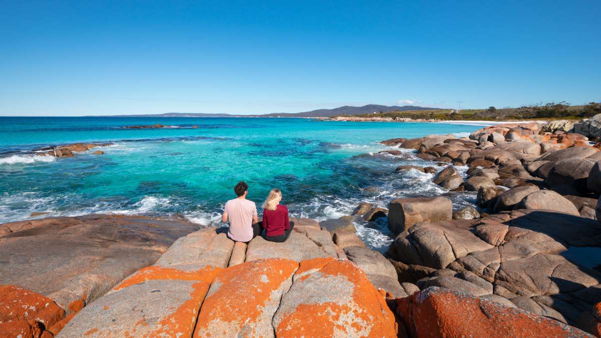 couple sitting on reddish rock overlooking clear blue ocean