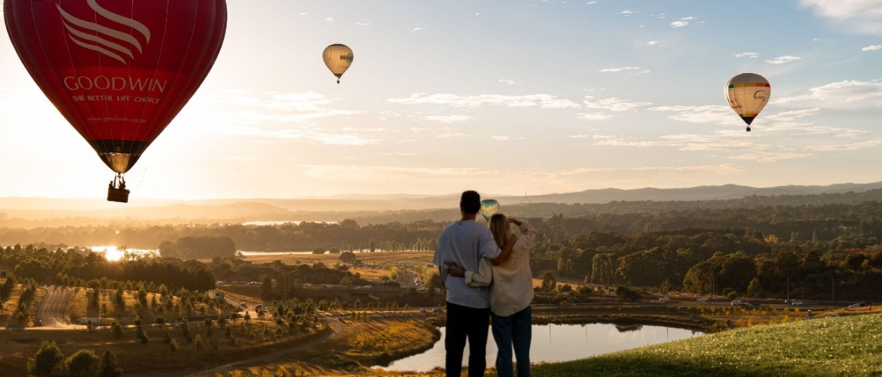 Two people with their arms around each other look out over scrivener dam from the National Arboretum, Canberra. It is sunrise and three hot air balloons can be seen in the sky