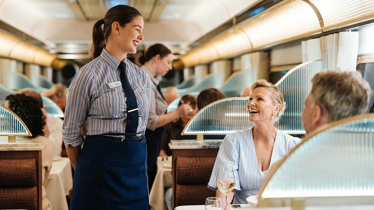 A hospitality worker smiles at two passengers enjoying the service in the Gold Premium Dining Carriage aboard The Ghan train in Australia.