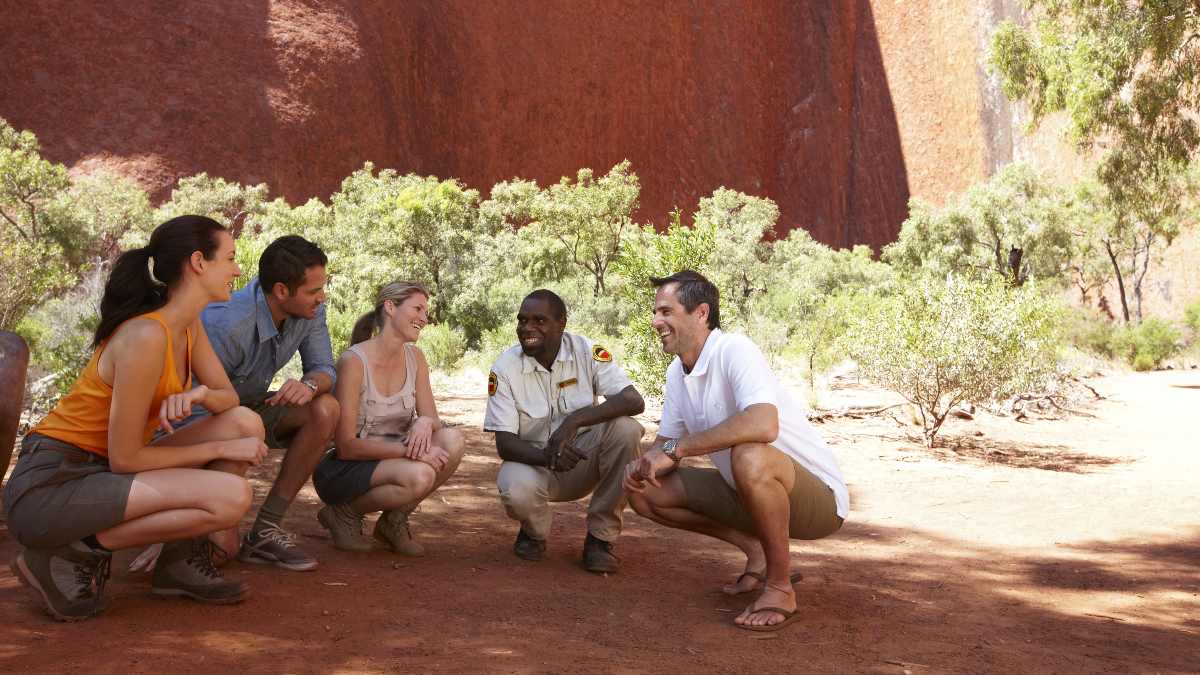 Indigenous Australian tour guide with four tourists crouched on ground at Uluru talking and smiling