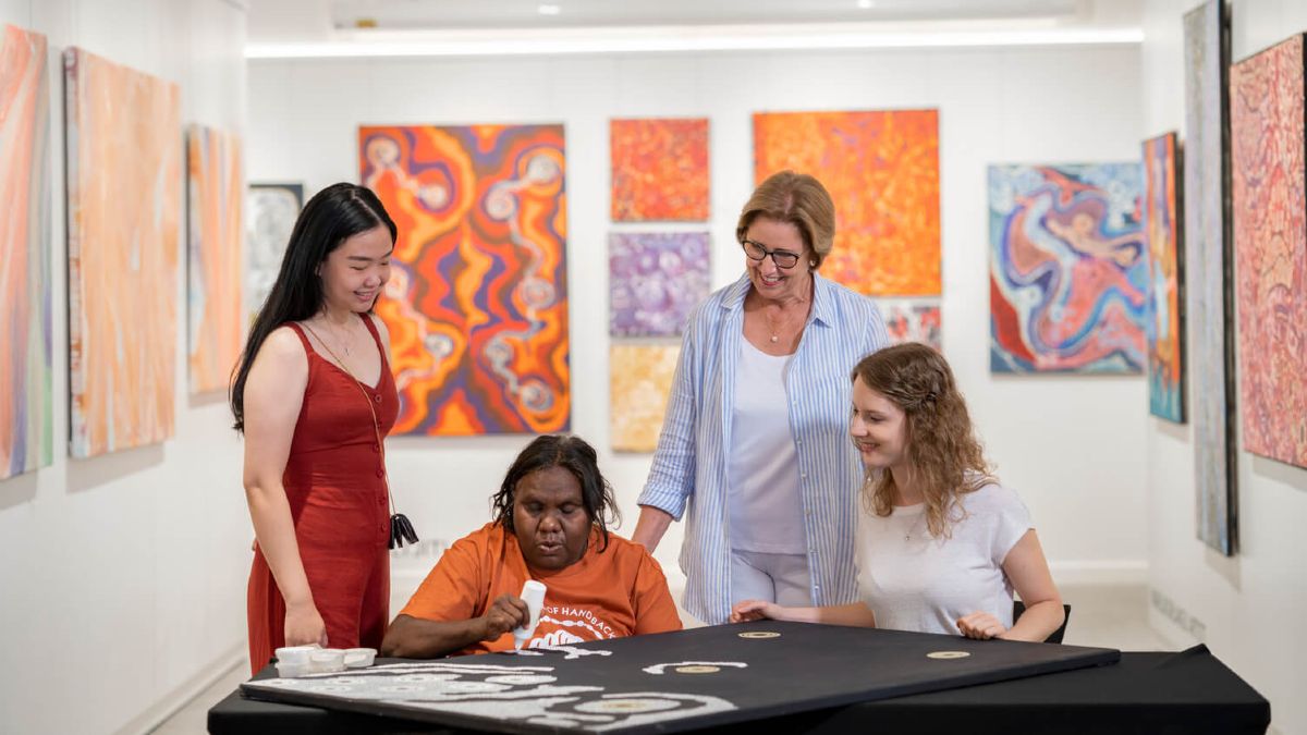 three tourists watch an Indigenous artist create artwork in a gallery