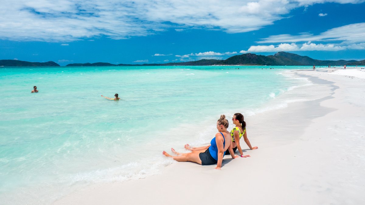 woman in swimsuits sitting on white sandy beach; two more people are floating in the ocean