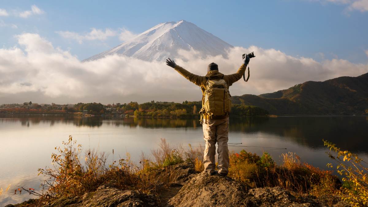 man standing on rock by lake looking toward Mt Fujiin Japan, holding arms aloft with a camera