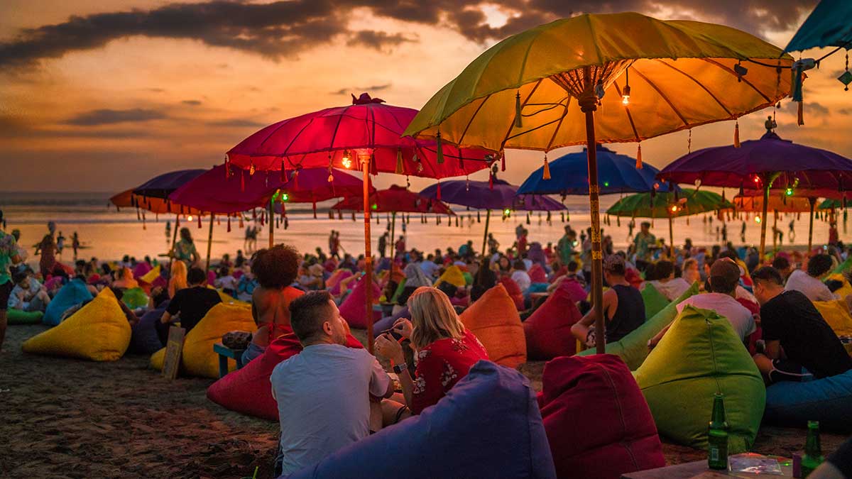 Tourists relax on colourful bean bags, under umbrellas, at sunset on beach in Denpasar, Bali, Indonesia.
