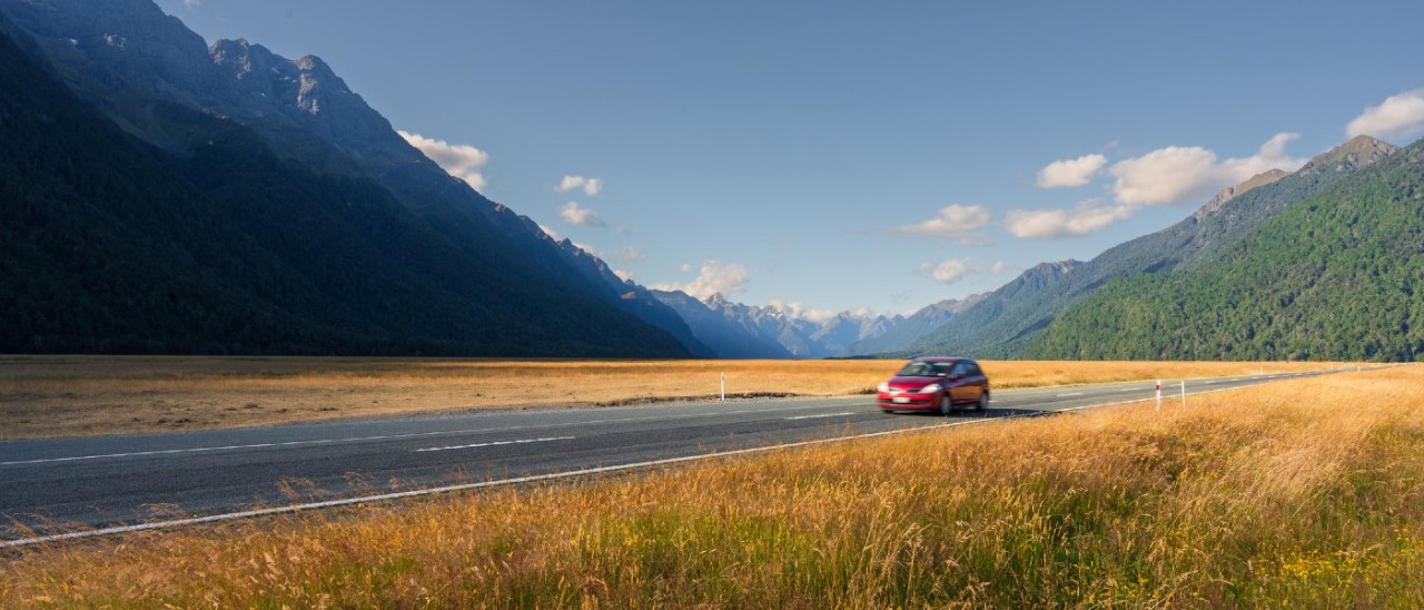 A small red hatchback car driving on a road approaching Milford Sound in New Zealand. The road is surrounded by golden plains of grass while tall mountains stand in the background