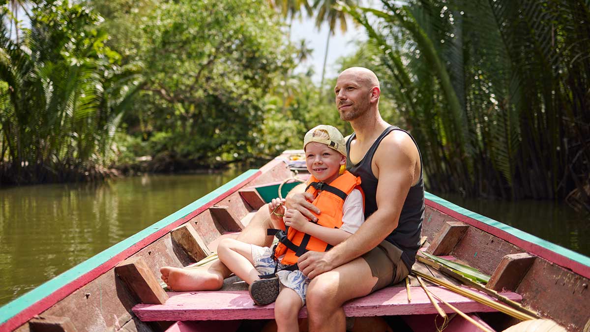 A man and school-age child wearing a life jacket sitting on a wooden long boat in a Thai canal